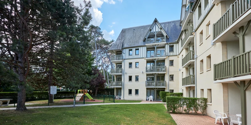 Apartment buildings stand tall with balconies, overlooking a lush, grassy courtyard featuring a small playground at Residence Les Embruns