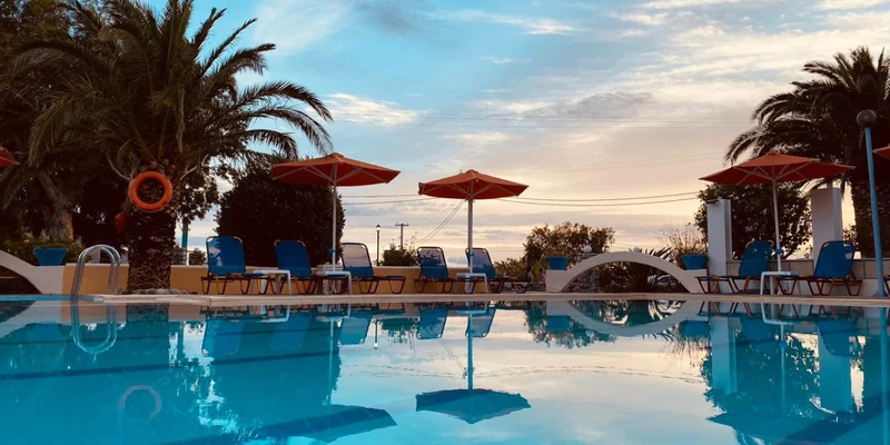 Pool reflecting blue sky and palm trees, surrounded by lounge chairs and umbrellas at Apollonia