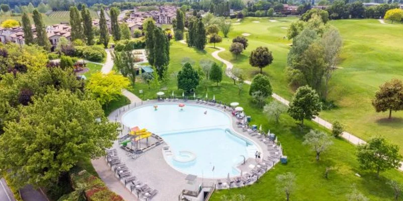 A large resort-style swimming pool with surrounding loungers in a landscaped park with paths, trees, and distant buildings at Residence Eden