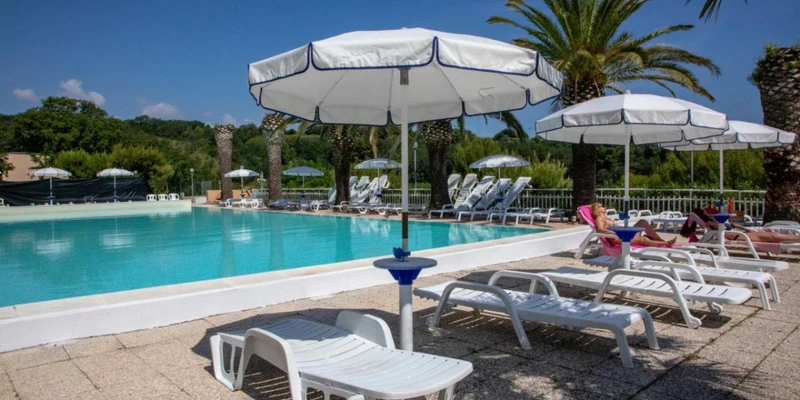 Lounge chairs with umbrellas beside a swimming pool surrounded by palm trees under a clear sky at Camping Village Mirage