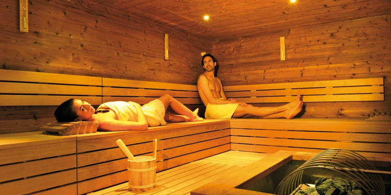 People relax on wooden benches in a warmly lit sauna; one lies down, the other sits upright at Seeblick Toni