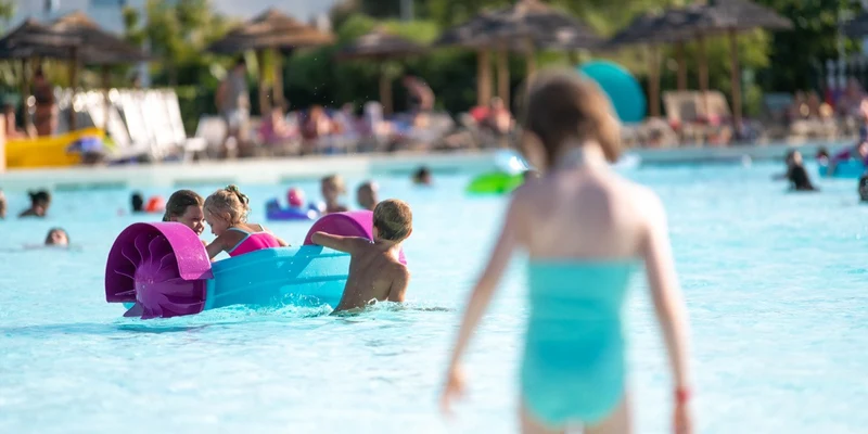 Children play with a floating watercraft in a swimming pool, with people relaxing and sun umbrellas in the background at I Turchesi Club Village