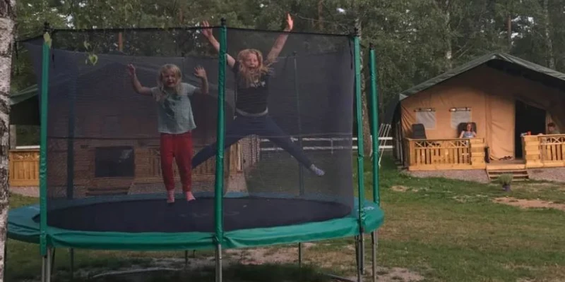 Two children jump on a trampoline in a forested campsite with tents and a wooden deck in the background at Smaland Miniglamping