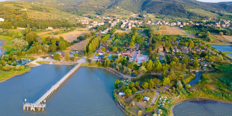 Pier extending over water, surrounded by trees and fields with small buildings populated nearby at Trasimeno Glamping Resort