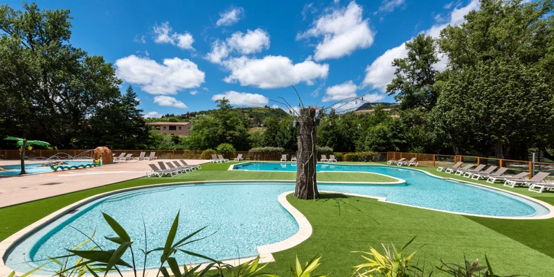 Outdoor pool surrounded by lounge chairs, a tree sculpture, and lush greenery under a bright blue sky at Le Millau Plage