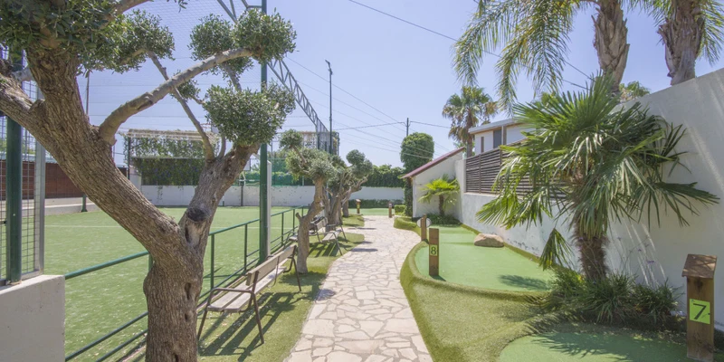 A stone path winds between trimmed trees and synthetic grass areas next to a green sports field at Alannia Els Prats