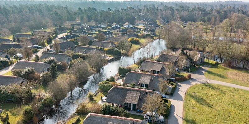 Thatch-roofed cottages line a narrow waterway surrounded by trees and greenery in a forested village setting at Vakantiepark de Veluwse Hoevegaerde