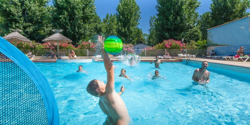 People playing with a ball in a swimming pool surrounded by lounge chairs and trees at Le Clos Virgile