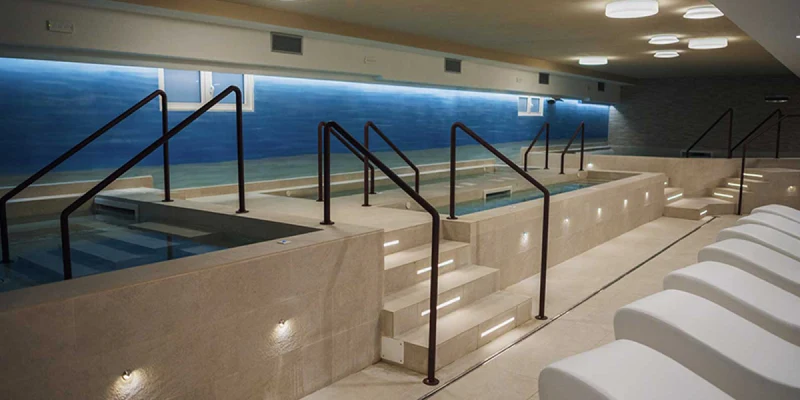 Indoor pool with steps and railings, surrounded by lounge chairs in a modern, well-lit spa setting at Villaggio Turistico Lido D Abruzzo