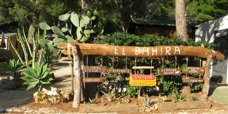 Wooden sign displaying "El Bahira" with direction markers surrounded by cacti and trees at a sunlit campsite at El Bahira