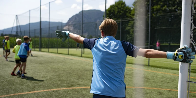 A goalpost goalkeeper wearing gloves directs players on a soccer field, with mountains and trees in the background at Continental Lido