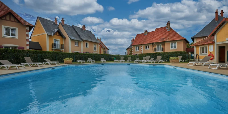 Swimming pool reflecting blue sky, surrounded by lounge chairs and colorful houses in a neatly landscaped area at Port Guillaume