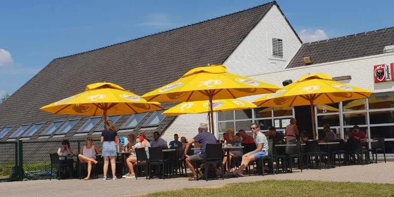 People sit and chat under yellow umbrellas outside a white building on a sunny day at Vakantiepark Breebos