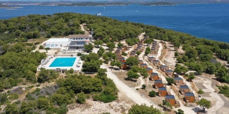 Aerial view of wooden cabins surrounded by trees, with a large swimming pool and buildings nearby, overlooking the sea at Obonjan Island Resort