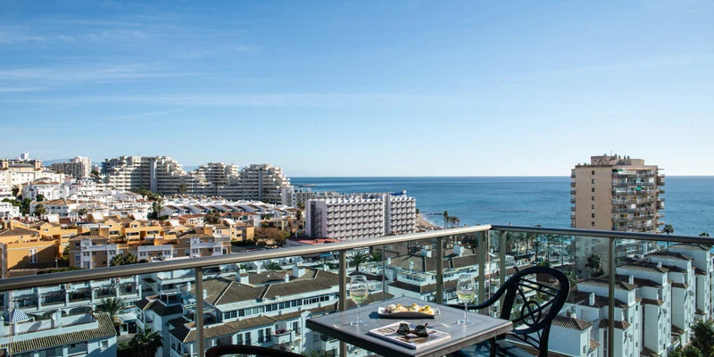 A balcony table holds two wine glasses, snacks, overlooking urban rooftops and a bright blue ocean at Benalmádena Príncipe