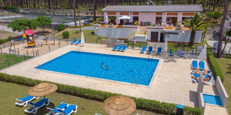 Swimming pool with people swimming, surrounded by lounge chairs and umbrellas, near a building and playground at Valado