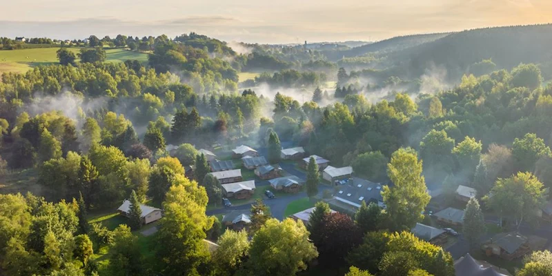 Aerial view of cabins nestled among trees with mist rising, surrounded by forests and hills at Landal Glamping Neufchâteau