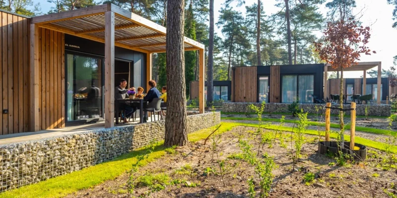 People dining at a wooden cabin terrace, surrounded by trees and grass in a forested campsite at Europarcs Hoge Kempen