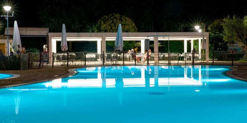 Illuminated swimming pool reflecting nearby lit dining area at night, surrounded by trees and umbrellas at Baia Verde
