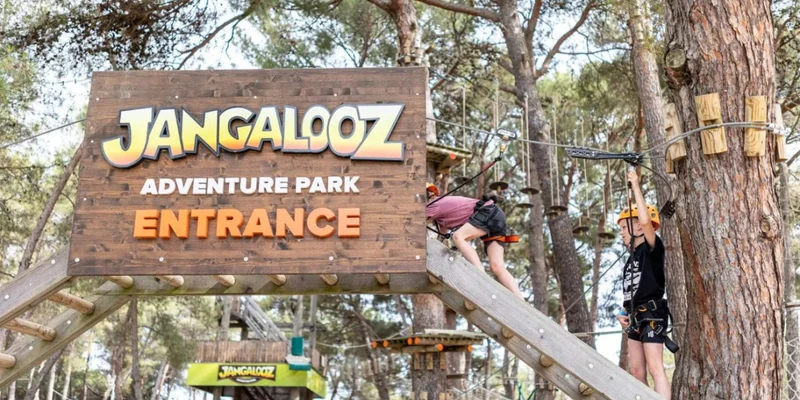 Sign reads "Jangaloz Adventure Park Entrance" with people climbing, surrounded by trees and adventure rope course structures at Bi-Village