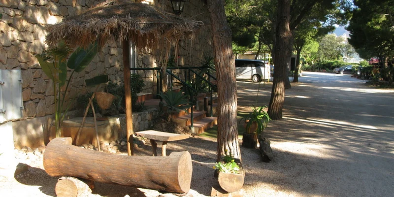 Wooden benches sit beneath a thatched umbrella outside a stone building, with trees providing shade and cars parked nearby at El Bahira
