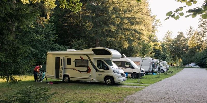 A person checks a bicycle next to a parked motorhome in a forested campsite at Forest camping Mozirje