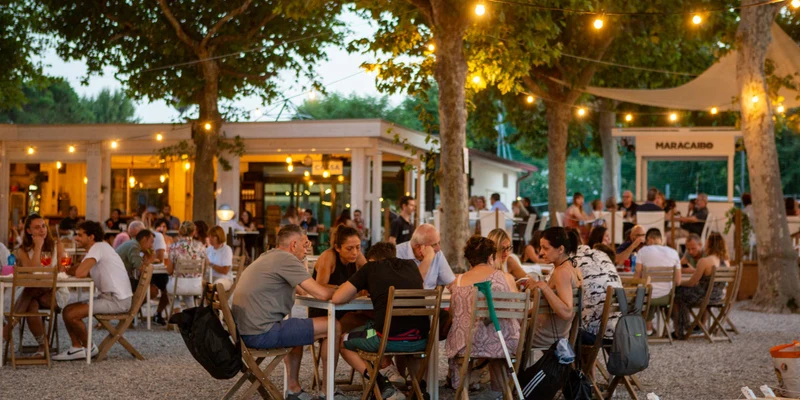 People seated at wooden tables, eating and socializing under string lights, surrounded by trees and outdoor restaurant setting at Del Sole