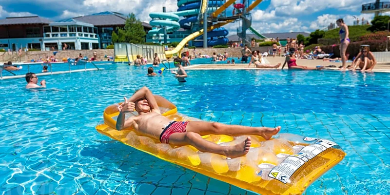 Child lounging on inflatable mattress, giving thumbs-up in busy outdoor swimming pool with water slides at Terme 3000