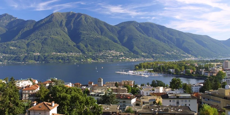 Buildings and lush greenery sit by a serene lake, with tall, green mountains and a blue sky in the background at Okay Lido