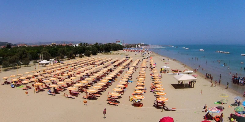 Rows of orange beach umbrellas line a sandy shore filled with sunbathers near a calm ocean at Riva Nuova