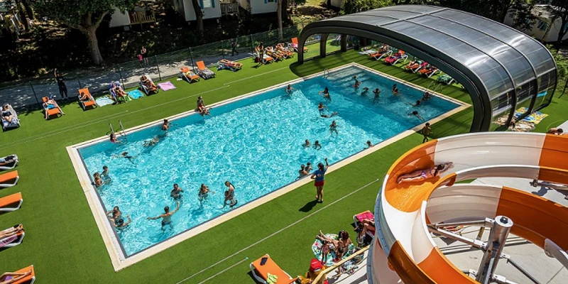 People swim in a rectangular pool near a covered area, surrounded by sunbathers on lounge chairs and a tall water slide at Les Ajoncs d'Or