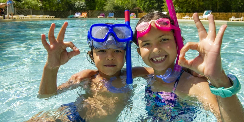Children wearing snorkel masks giving an 'OK' sign while swimming in a pool, surrounded by trees and lounging chairs at Aqua Viva