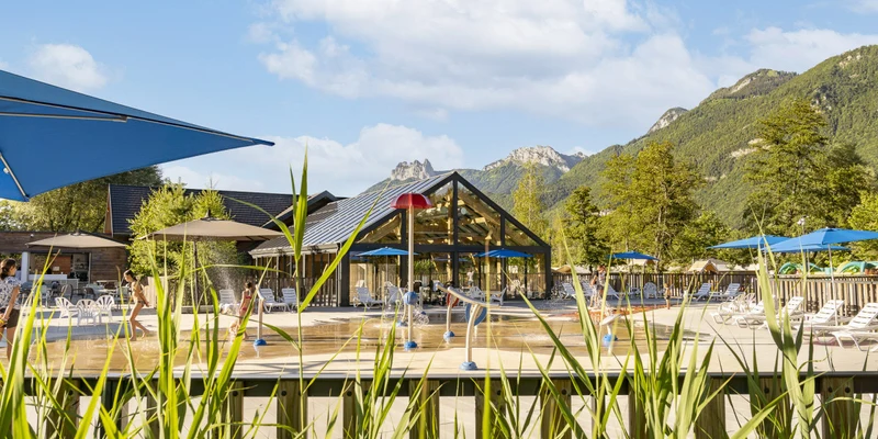 Children playing in a pool with fountains, surrounded by lounge chairs and a glass building, mountains in the background at La Nublière