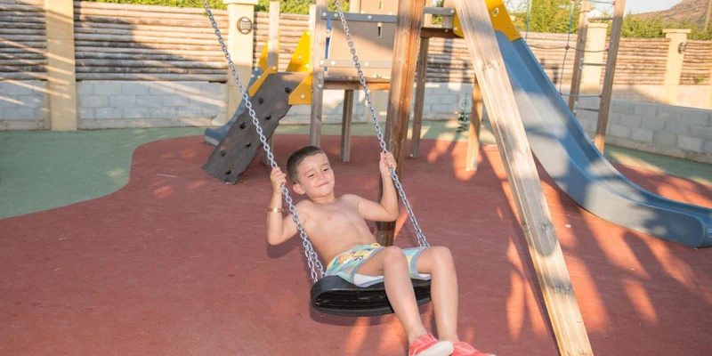 Child swinging on a swing set, in a playground with a slide and climbing structure in the background at La Prairie 