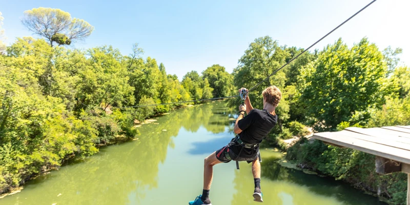 Person ziplining over a river surrounded by lush greenery on a sunny day at Marvilla Parks Le Domaine de Massereau