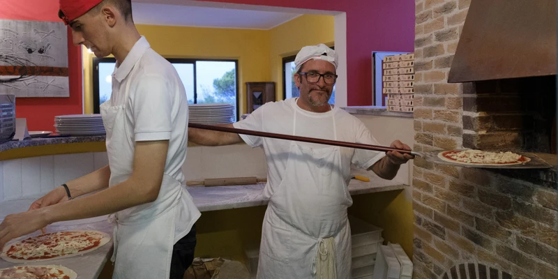 Pizzaiolo placing pizza into brick oven, another person preparing pizzas in a colorful kitchen at Mar Y Sierra
