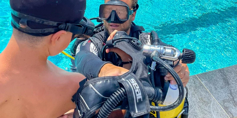 Scuba instructor assisting a young boy near a pool, both wearing diving masks; water shimmers in sunlight at Baia Blu La Tortuga