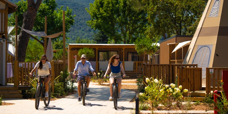 Three people ride bicycles on a sunny path, surrounded by wooden cabins and trees at Trasimeno Glamping Resort