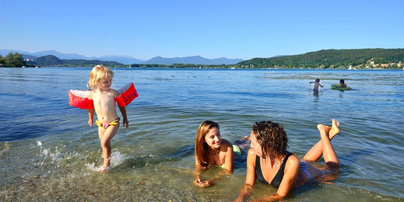 Child walks at water's edge, while two women lie in shallow water; lake, distant hills, other swimmers in background at Eden Maggiore