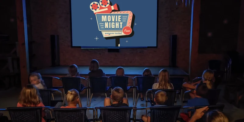 Children and adults watching a movie on a large screen inside a dark hall with bunting decorations at Chvalsiny