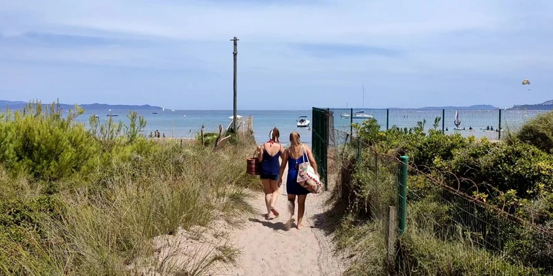 Two women walking on a sandy trail towards a beach with boats and distant mountains at La Bergerie Plage