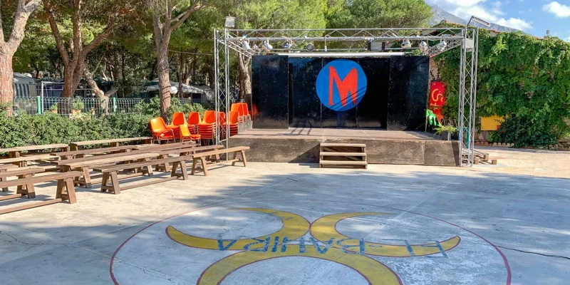 Outdoor stage with wooden benches and stacked chairs in a wooded campground at El Bahira