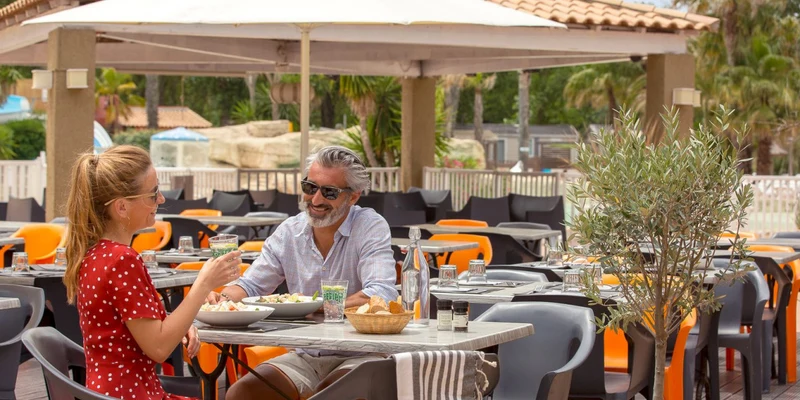 A man and woman enjoying a meal outdoors on a shaded patio with tables and chairs surrounding them at Les Vagues