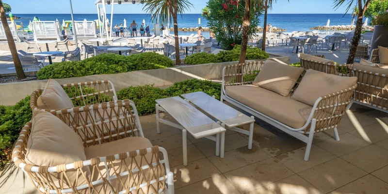 Outdoor seating area with wicker chairs and tables, overlooking a beachfront scene with umbrellas and the sea at Aminess Maravea Camping Resort