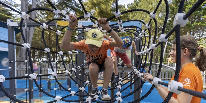 Children climbing rope tunnel at playground, woman supervising nearby, surrounded by trees and outdoor setting at Moulin de l'Eclis