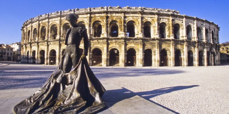 A statue of a matador stands before a large Roman-style amphitheater under a clear blue sky at La Clémentine