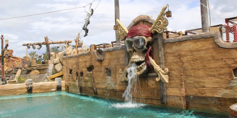 A pirate-themed zip-liner gliding above a wooden ship with a skull fountain into a pool of water at Resort Cap Pirate