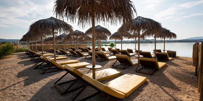 Wooden lounge chairs line a sandy lakeside under straw umbrellas, overlooking calm water at Amadria Park Camping Sibenik