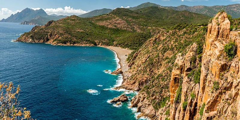Cliffs overlook turquoise sea, waves crashing against rocks, and a curving sandy beach under a partly cloudy sky at Torraccia