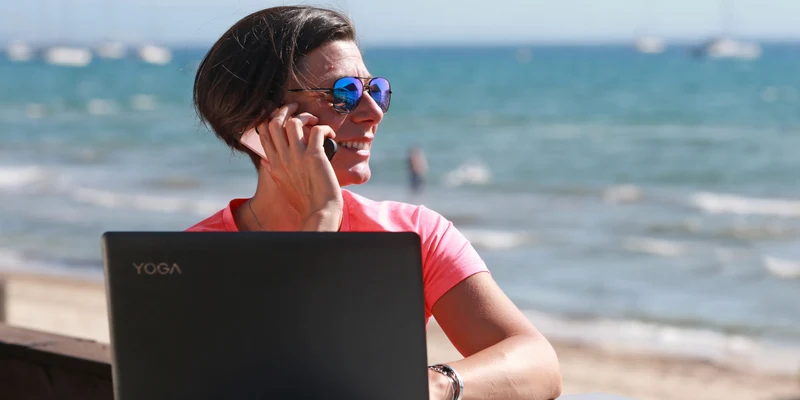 Woman talking on phone, using laptop at a beachside table with boats in the distance at Eurosurf